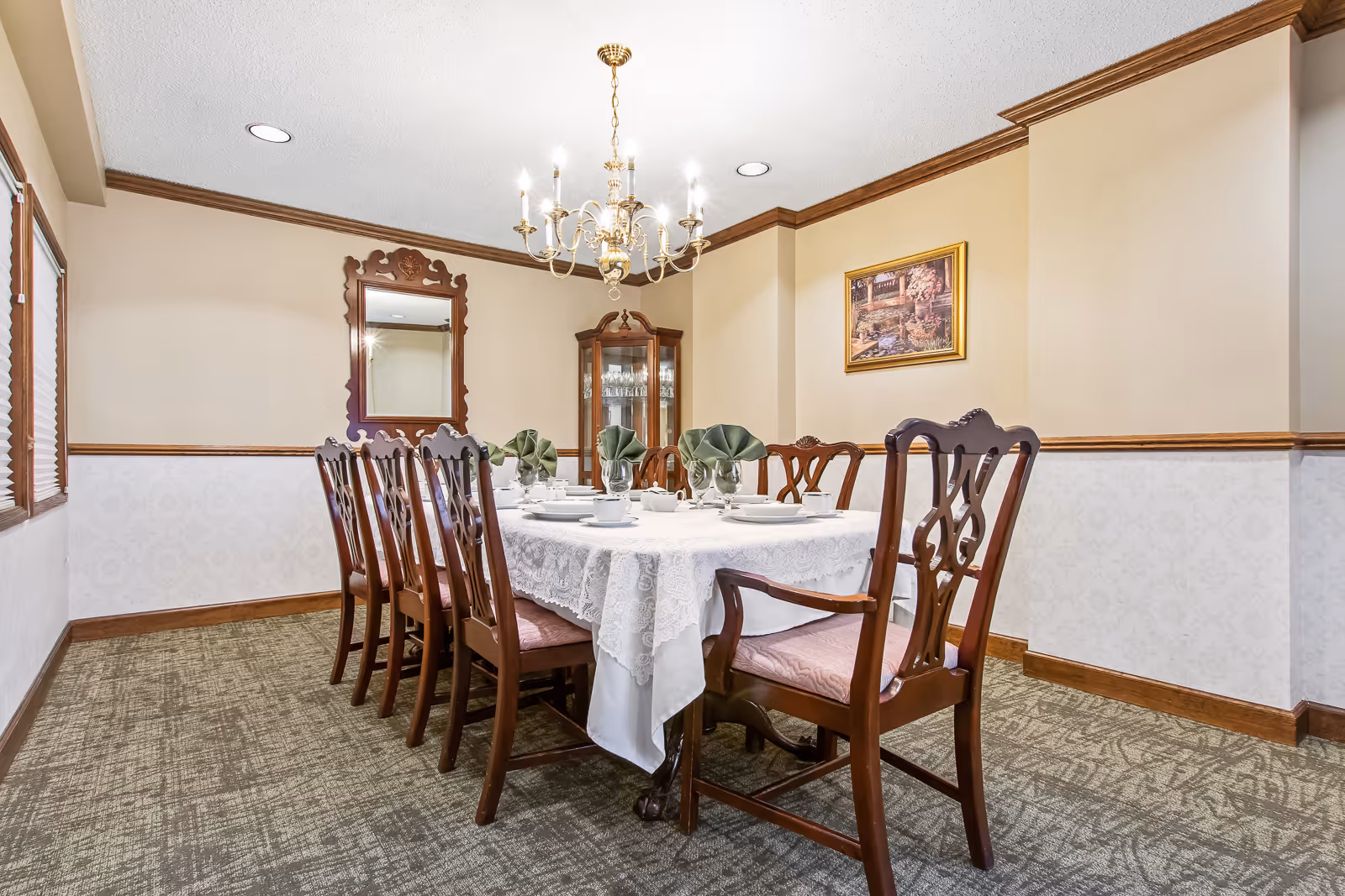 A formal dining room with a long wooden table covered with a white lace tablecloth, set with white dishes, cups, and green folded napkins in glasses. The room features eight wooden chairs with pink cushions, a chandelier hanging from the ceiling, a decorative mirror on one wall, a glass-fronted cabinet with glassware, and a framed painting on the wall. The walls are beige with wooden trim, and the floor is carpeted.