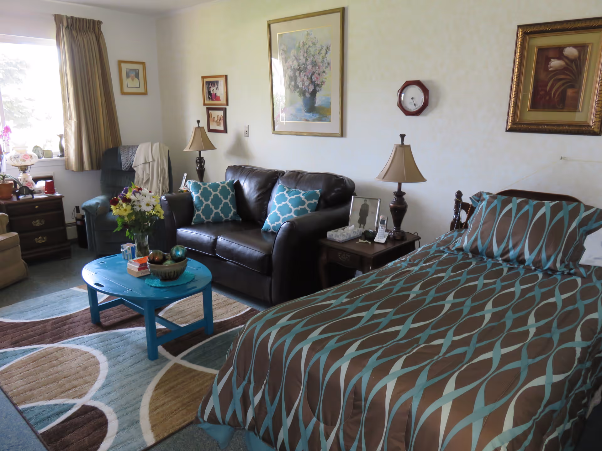 A cozy senior living room and bedroom combined space featuring a bed with a brown and turquoise patterned comforter, a dark brown leather loveseat with two turquoise patterned pillows, a blue coffee table with decorative items, a side table with a lamp and phone, a recliner chair near a window with beige curtains, and framed artwork on the walls.