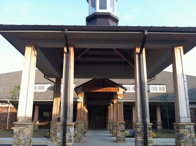 Entrance of a building with a covered walkway supported by wooden pillars with stone bases, leading to a door. The building has a brick exterior and a gray roof with a small tower structure on top.