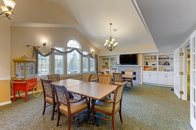 A spacious, well-lit common room with multiple tables and chairs arranged for group seating. Large windows with decorative curtains allow natural light to fill the room. A popcorn machine is positioned in the corner. The far wall features built-in white shelves and cabinets with a flat-screen TV mounted above a fireplace. The room has a carpeted floor and a chandelier hanging from the ceiling.