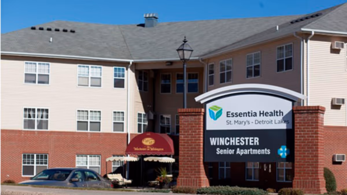 Front exterior of a multi-story senior apartment building with a large sign reading Essentia Health Winchester Senior Apartments and a parked car.
