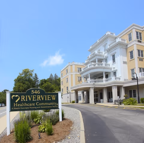 Exterior view of Riverview Healthcare Community building with a multi-story structure featuring balconies and columns. A green sign in front reads '546 Riverview Healthcare Community, A Health Concepts Nursing and Rehabilitation Facility.' The sky is clear and blue with some greenery around the sign.