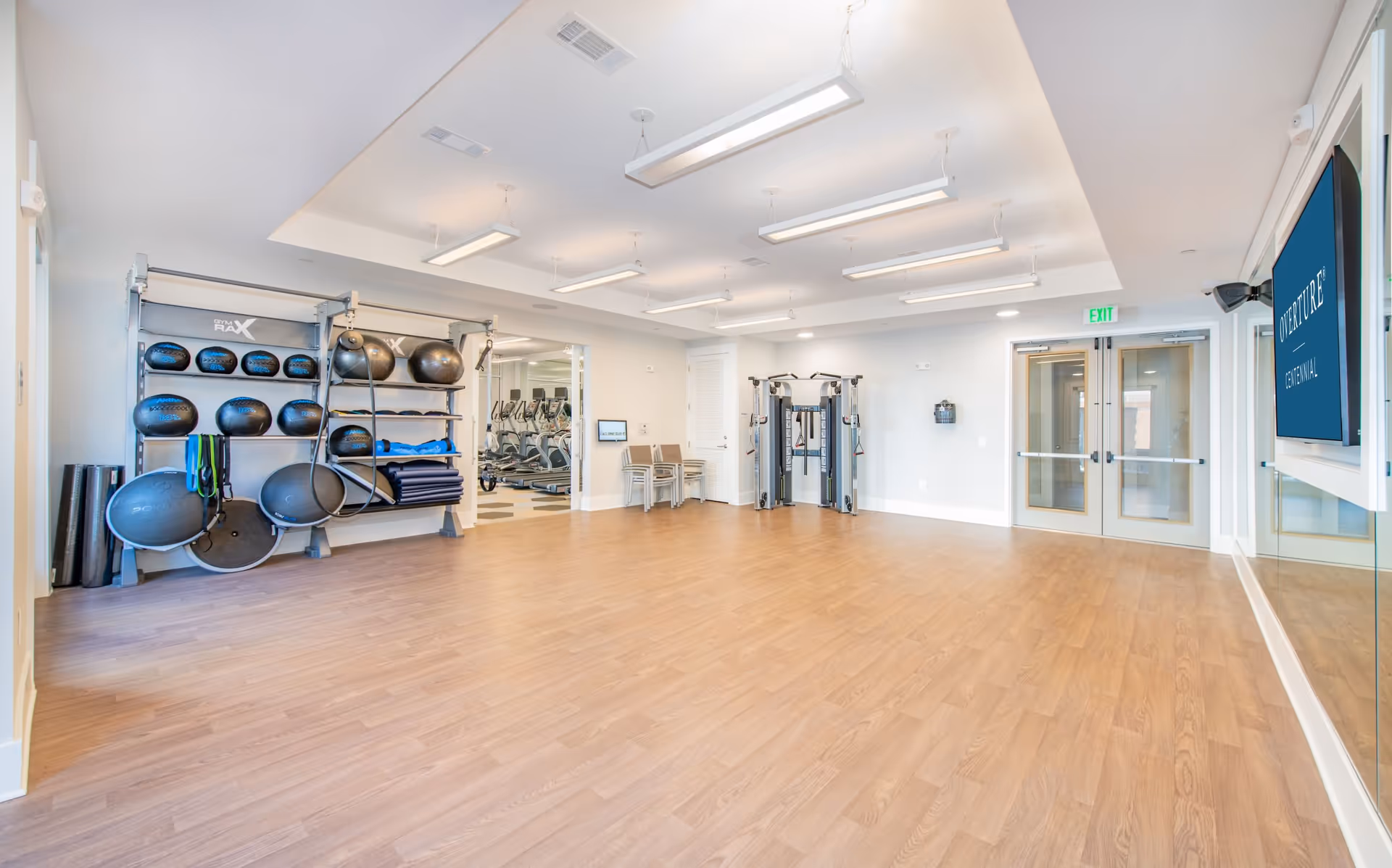 Bright fitness studio with wood flooring, exercise equipment and storage racks along one wall and double doors at the far end.