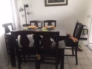 A dining area with a dark wooden table and six matching chairs. The table is set with placemats, plates, and some food items. There is a floor lamp with three lights in the corner near a window with light-colored curtains. The floor is tiled in white.