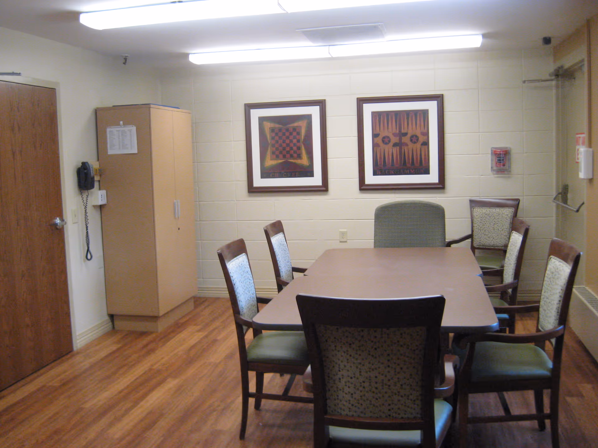 Rectangular table surrounded by chairs in a small common room with framed artwork, a storage cabinet, and wood-look flooring.