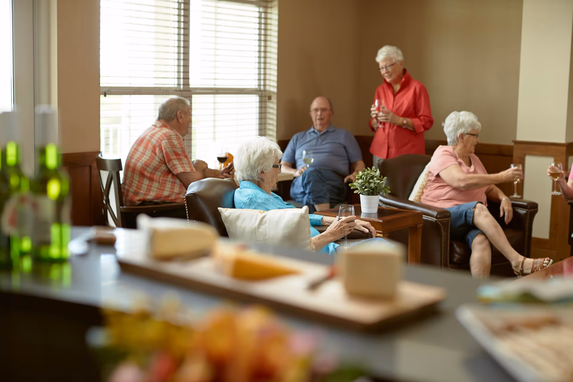 A group of elderly people socializing in a cozy living room area with large windows and wooden trim. Some are seated on leather chairs holding glasses of wine, while one woman stands holding a glass. A small table with a potted plant is between the chairs, and a blurred foreground shows bottles and food on a counter.