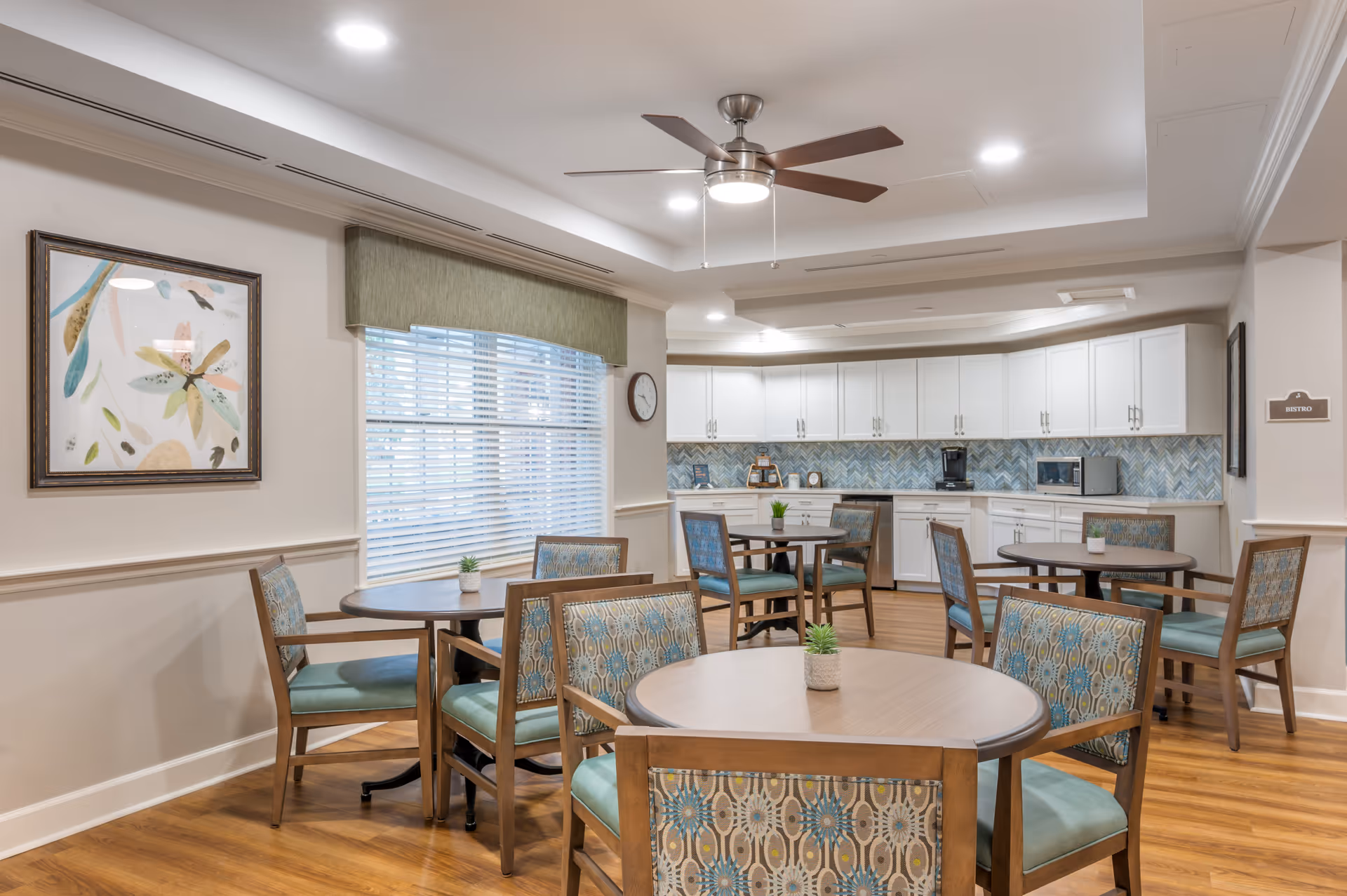 A bright and clean dining area with several round wooden tables and patterned cushioned chairs. The room features a ceiling fan with lights, a large window with blinds and a green valance, and a kitchen area with white cabinets, a blue patterned backsplash, and appliances including a coffee maker and microwave. A framed abstract artwork hangs on the wall.