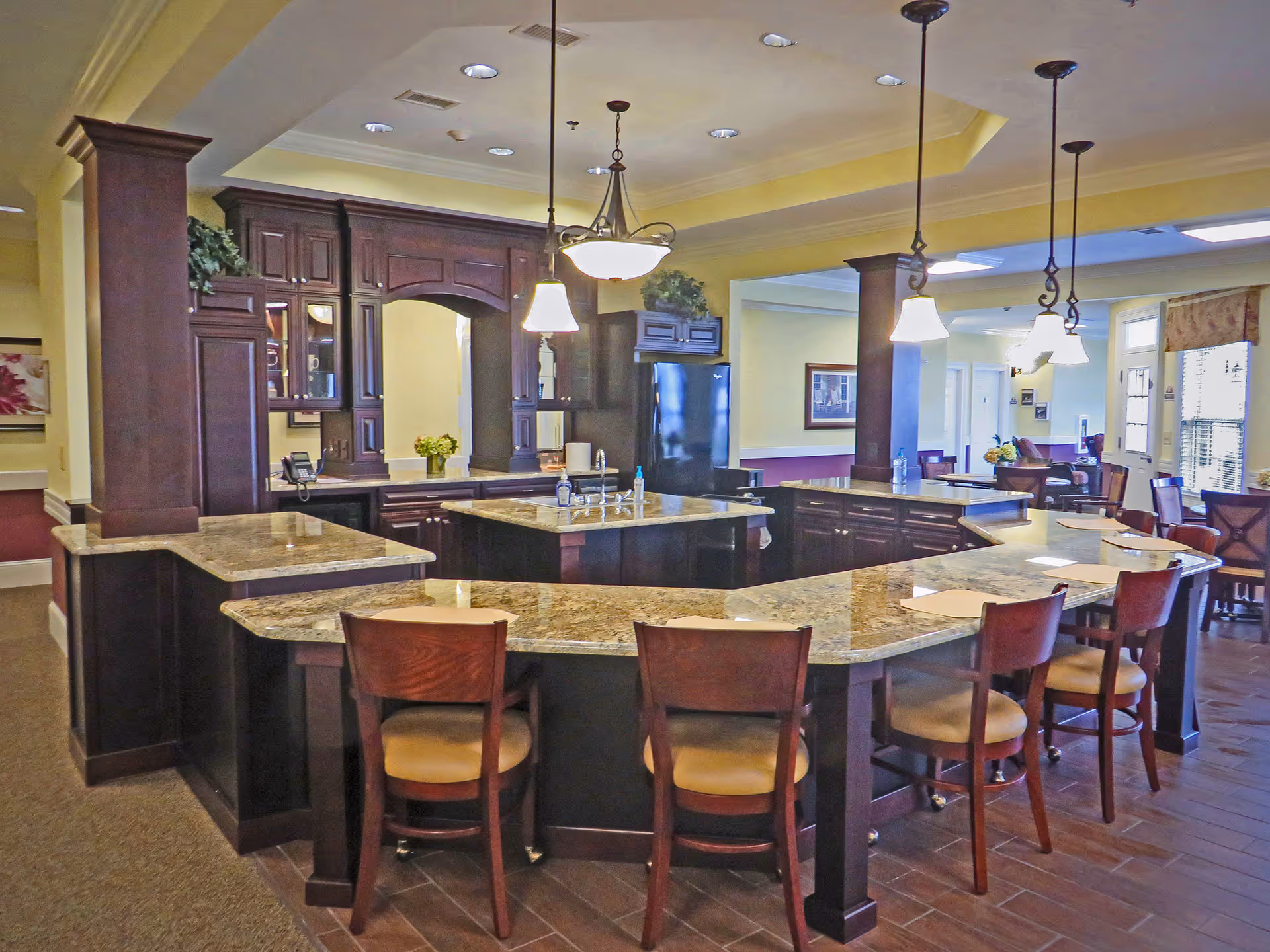 Interior view of a senior living facility kitchen area with a large granite countertop island surrounded by wooden chairs. The kitchen features dark wood cabinetry, pendant lighting, and a refrigerator. The space opens into a common area with additional seating and windows allowing natural light.