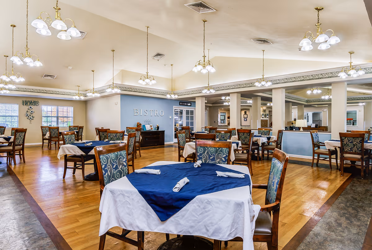 A spacious dining room with multiple tables covered with white and blue tablecloths, each set with rolled napkins. The room features wooden floors, patterned upholstered chairs, and multiple hanging light fixtures. A blue wall with the word 'BISTRO' is visible in the background along with windows letting in natural light.