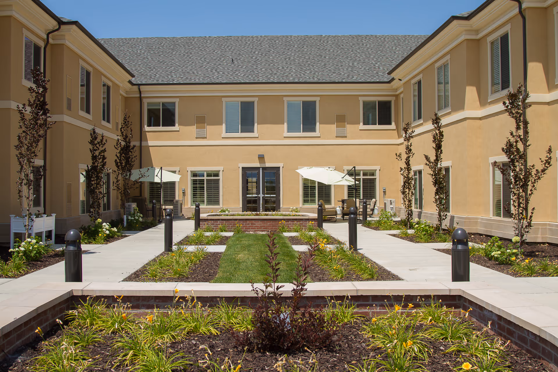 Outdoor courtyard area of a senior living facility with a central garden bed featuring plants and flowers, surrounded by paved walkways and small trees. The beige two-story building with multiple windows encloses the courtyard, and there are patio umbrellas and seating areas near the building walls.