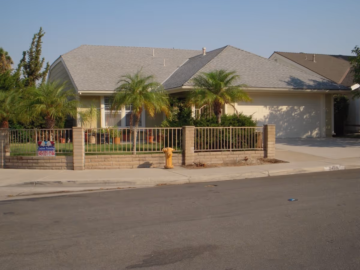 Single-story residential house with a gray shingled roof, beige exterior walls, and a two-car garage. The front yard is enclosed by a low brick and metal fence, featuring several palm trees and green shrubs. A yellow fire hydrant is visible on the sidewalk in front of the house. The sky is clear and blue.