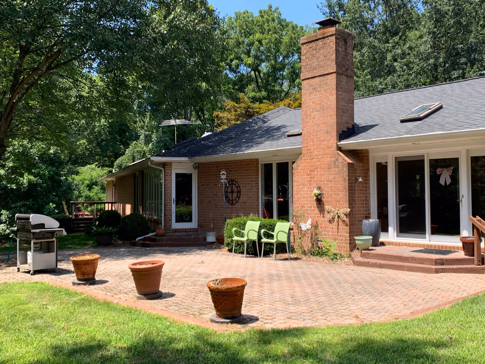 Back patio of a brick single-story building with a chimney, sliding glass doors, green chairs, potted planters and a gas grill.