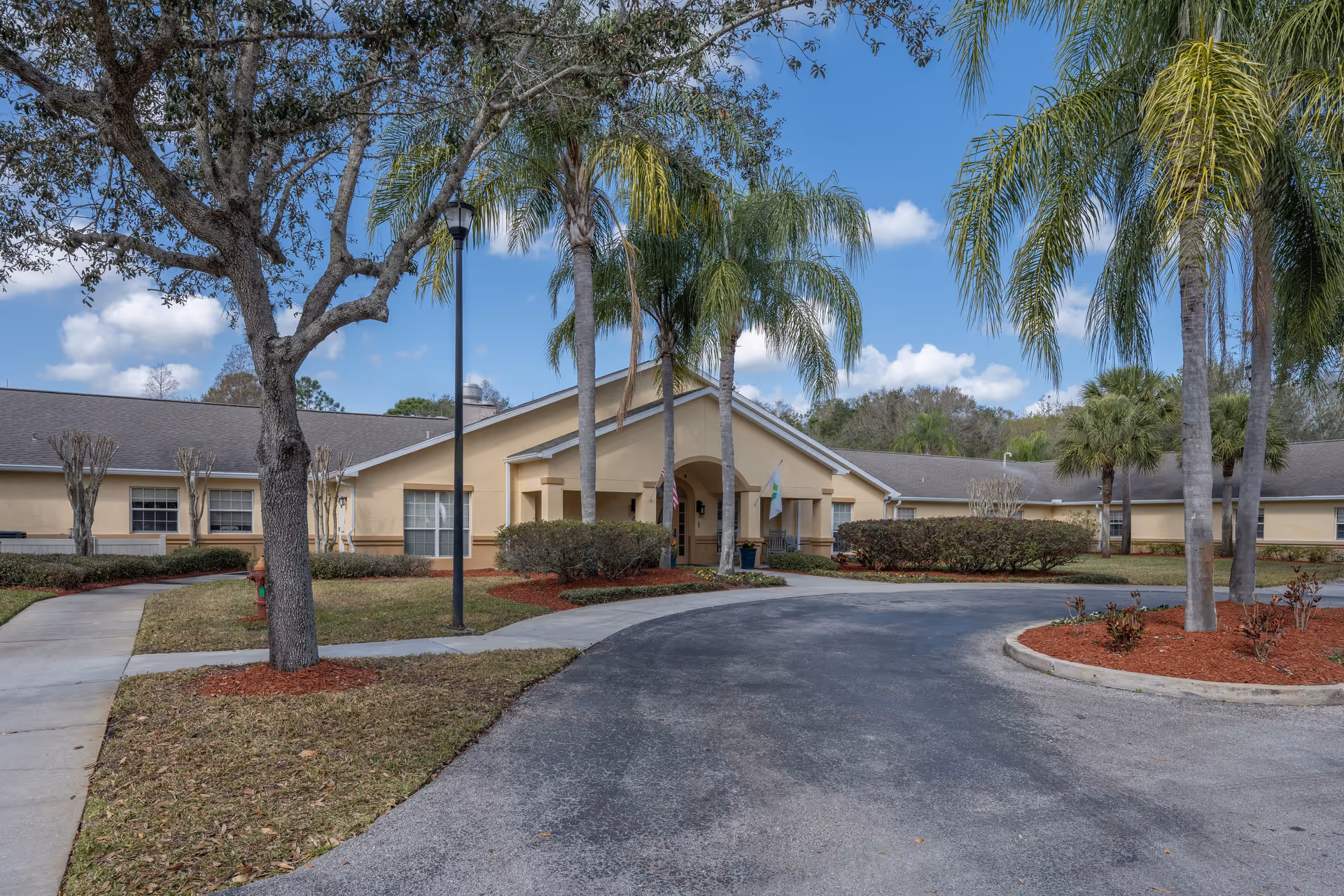 Exterior view of a single-story beige building with a peaked roof, surrounded by palm trees and landscaped bushes. A curved driveway leads to the entrance, which has a covered porch with two flags displayed. The sky is blue with scattered clouds.