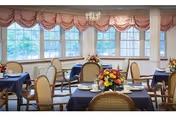 A dining room with several round tables covered with dark blue tablecloths, each set with white plates and silverware. The chairs have light-colored cushions and wooden frames. Large windows with pink valances allow natural light to fill the room. Floral centerpieces decorate each table.