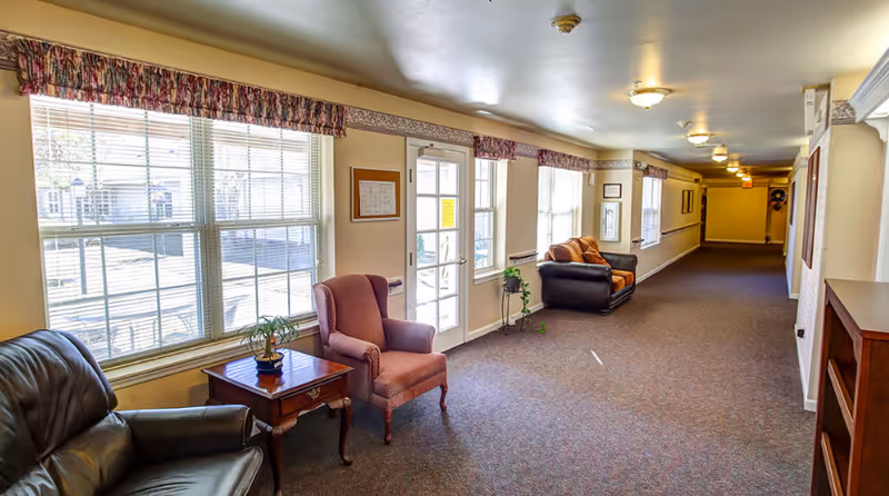 A long, well-lit interior hallway with large windows and seating, featuring armchairs, side tables, and potted plants along a carpeted corridor.