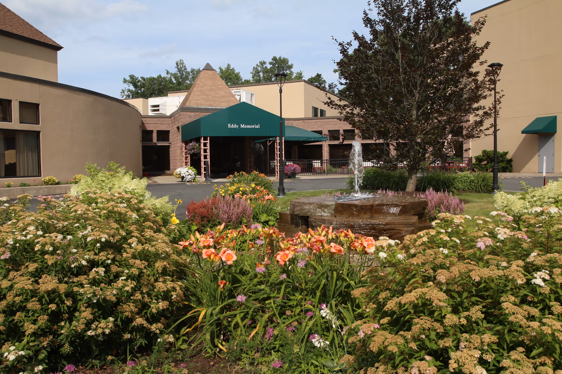 Entrance to Eddy Memorial building with a green awning, surrounded by a garden with colorful flowers and a small water fountain in front. The building has beige and brown walls with windows and a tree near the fountain.