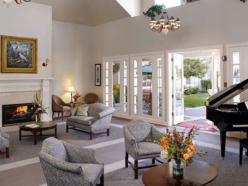 A bright and spacious living room with comfortable upholstered chairs and a loveseat arranged around a coffee table with a floral centerpiece. There is a fireplace with a painting above it on the left wall, a chandelier hanging from the ceiling, and large French doors open to a green outdoor patio area. A grand piano is positioned on the right side of the room.