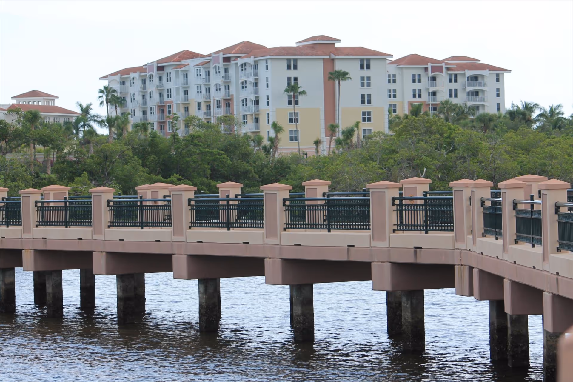 A large multi-story residential building with balconies and red-tiled roofs is visible behind a dense area of green trees. In the foreground, there is a wide pedestrian bridge with beige pillars and black railings extending over a body of water.