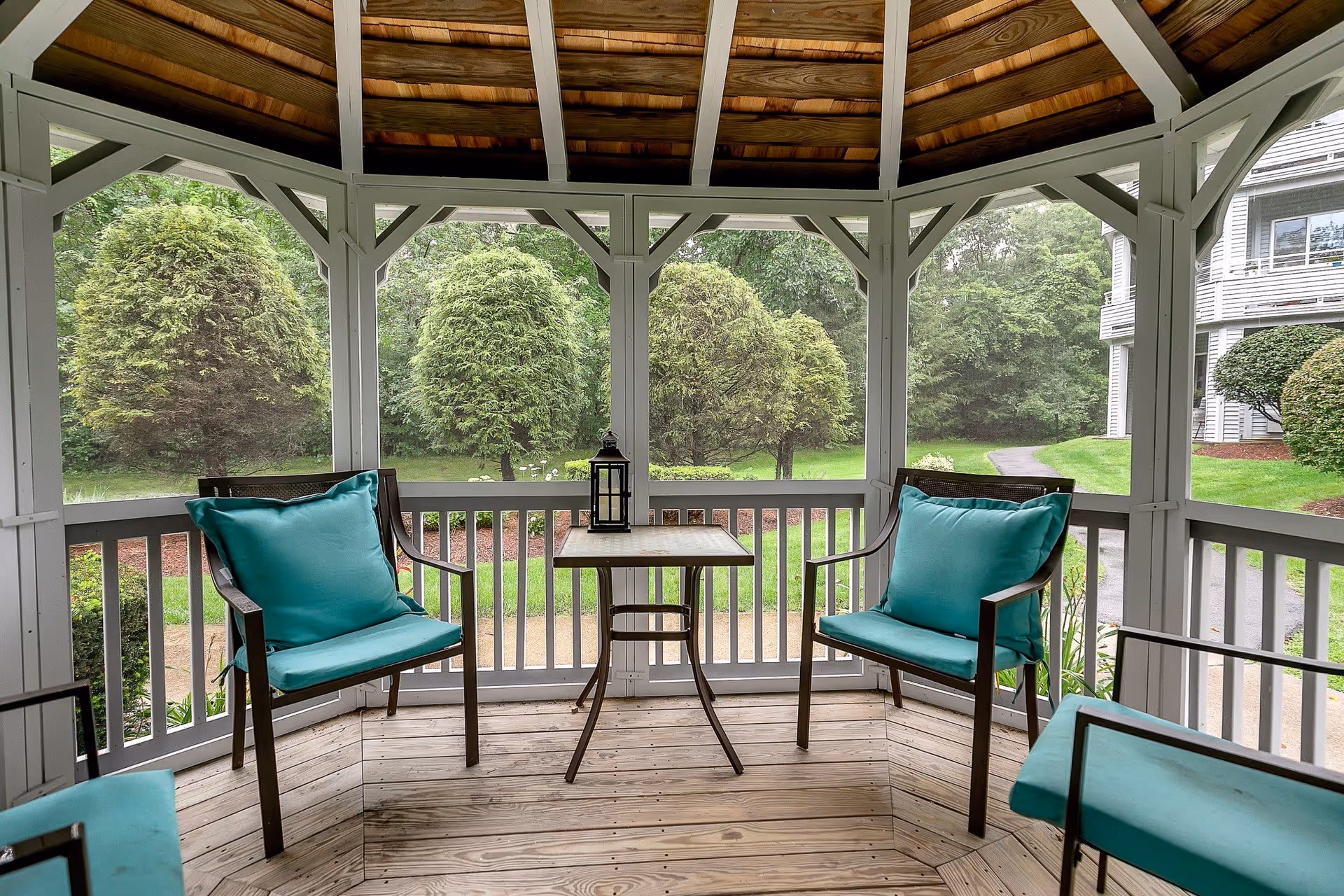 View from inside a wooden gazebo with a wooden floor and ceiling, furnished with metal chairs that have teal cushions and a small square table with a lantern on top. The gazebo overlooks a green lawn with neatly trimmed bushes and trees, and a paved pathway leading to a building in the background.