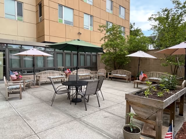 Outdoor patio area at a senior living facility with several wooden benches, tables with umbrellas, potted plants, and small American flags. The patio is surrounded by a building and some greenery.