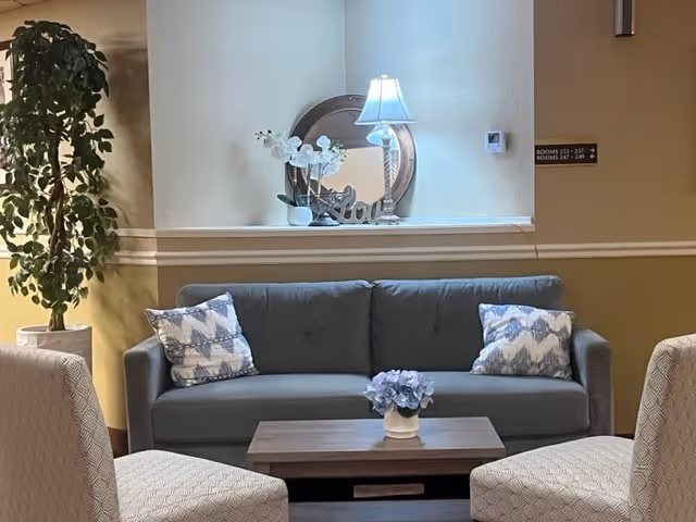 Seating area with a gray sofa flanked by patterned pillows, two upholstered chairs, a coffee table with flowers, and a lamp with a decorative mirror on a shelf behind.
