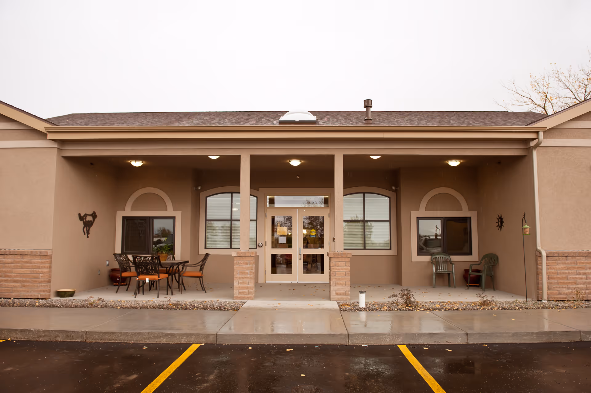 Exterior view of the entrance to Colorow Care Center, featuring a covered porch area with outdoor seating including tables and chairs. The building has beige walls with brick accents and large windows on either side of the double glass doors. The ground is wet, indicating recent rain, and there are parking spaces in front.