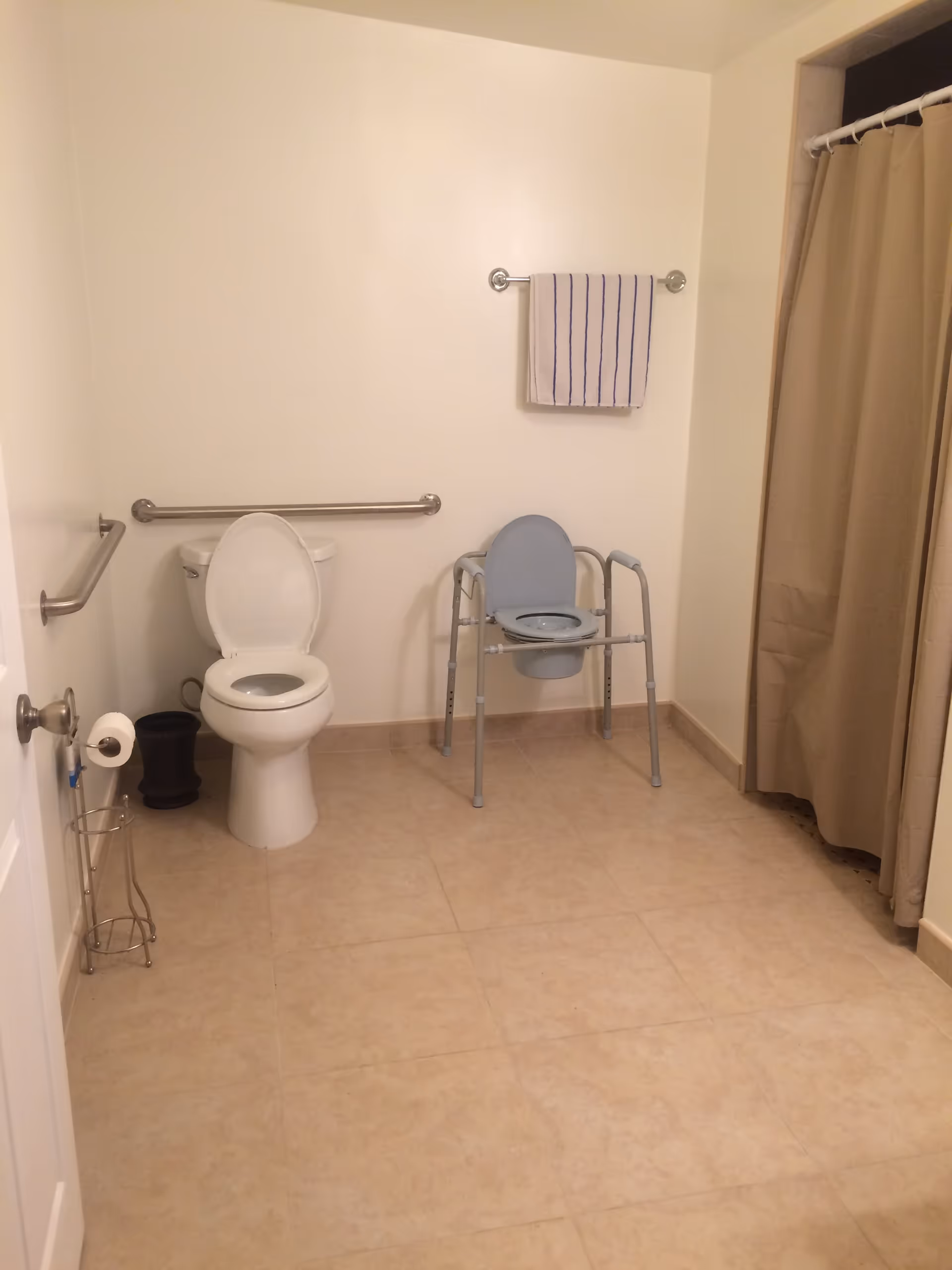 A clean and spacious bathroom with beige tiled floor and white walls. The bathroom features a white toilet with the seat lid open, a metal grab bar on the wall behind the toilet, and another grab bar on the adjacent wall. There is a towel rack with a white towel with blue stripes hanging on it. Next to the toilet is a metal stand holding a roll of toilet paper and a small black trash bin. To the right, there is a beige shower curtain partially covering a shower area. In front of the toilet, there is a gray commode chair with armrests and a removable seat.