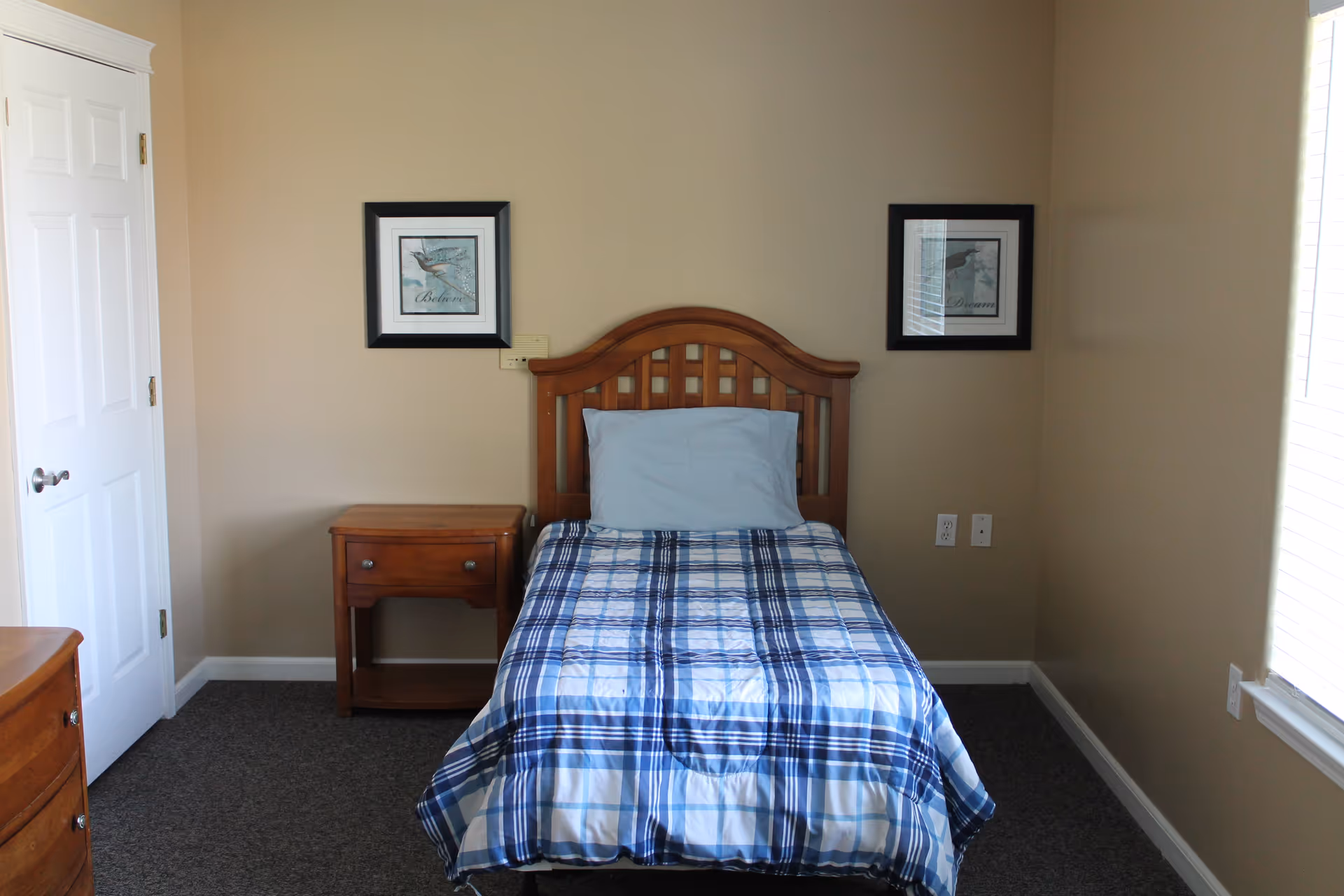 A small bedroom with a single wooden bed featuring a blue and white plaid bedspread and a light blue pillow. There is a wooden nightstand with a drawer next to the bed, two framed pictures hanging on the beige wall above the bed, a white door to the left, and a window with blinds on the right side letting in natural light.