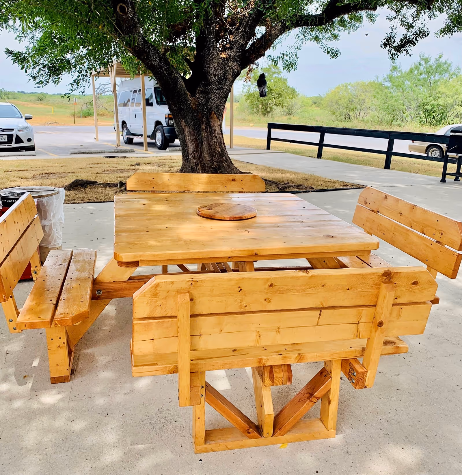 Outdoor wooden picnic table with attached benches under a large tree, with a parking lot and vehicles visible in the background.