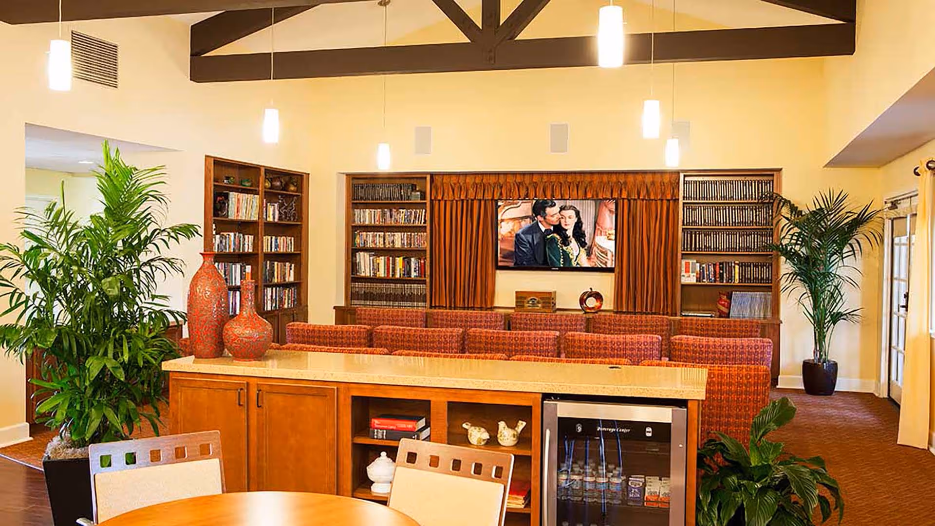 A cozy living room area in a senior living facility with a large flat-screen TV mounted on the wall, flanked by bookshelves filled with books and DVDs. There are multiple red patterned chairs arranged in rows facing the TV. The room features warm lighting with hanging pendant lights, wooden beams on the ceiling, and several green potted plants. In the foreground, there is a countertop with decorative red vases and a small refrigerator stocked with beverages underneath.