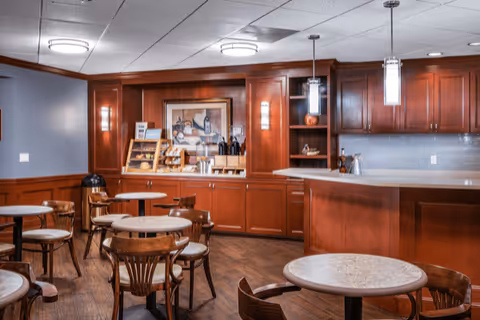 A cozy dining area with round tables and wooden chairs, featuring warm wooden cabinetry and a countertop with pendant lights hanging above. The back wall has a framed artwork and a small display shelf with various items.