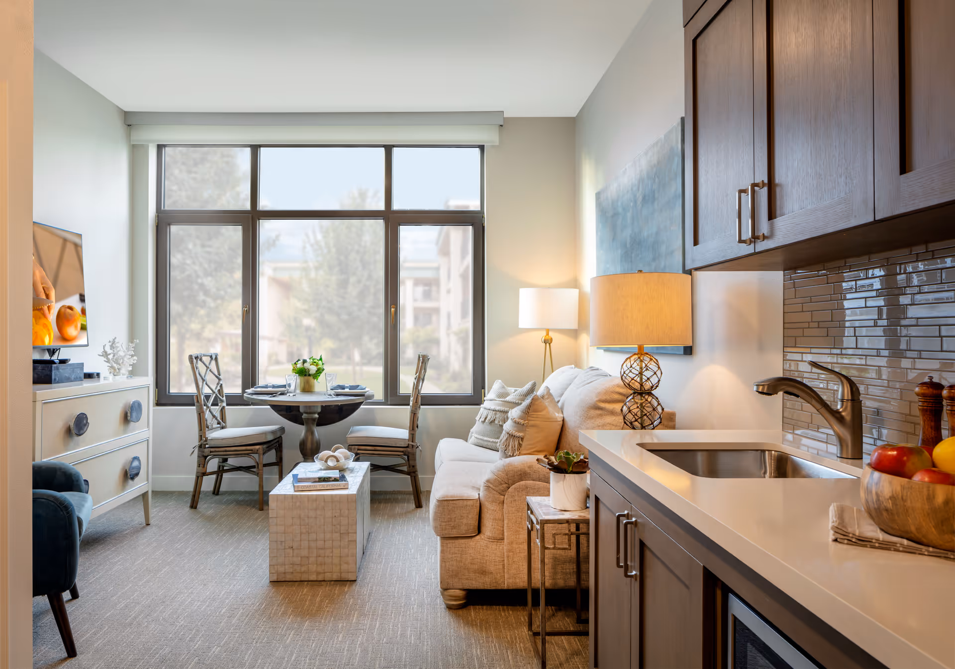 A bright and cozy living area in a senior living facility featuring a beige sofa with cushions, a small round dining table with two chairs near large windows, a flat-screen TV on a cabinet, and a kitchenette with a sink, countertop, and wooden cabinets. A table lamp and decorative plants add warmth to the space.