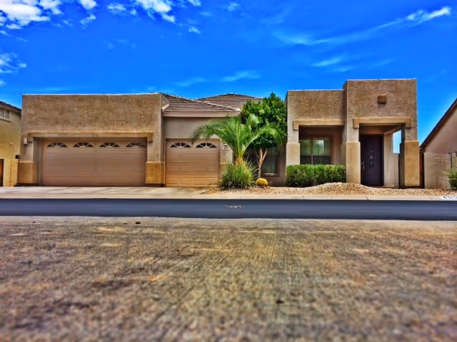 Front exterior view of a single-story desert-style home with a three-car garage, a small palm tree, and desert landscaping under a bright blue sky with some clouds.