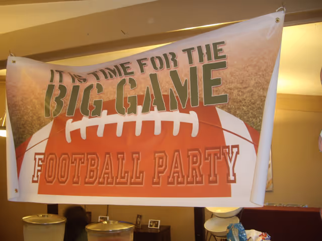 A banner hanging indoors with the text 'IT IS TIME FOR THE BIG GAME FOOTBALL PARTY' featuring a large football graphic in the background. Below the banner, part of a room is visible with containers and some furniture.
