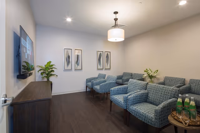 A waiting room with multiple blue patterned armchairs arranged along the walls. A flat-screen TV is mounted on the left wall above a dark wooden cabinet. Three framed feather artworks hang on the far wall. A round side table with bottled drinks and a small plant is on the right. The room has wood flooring, white walls, and a ceiling light fixture.