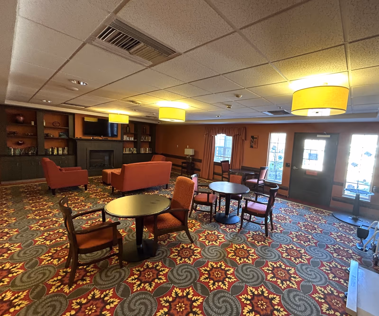 A cozy common area in a senior living facility with patterned carpet, several round tables with chairs, and a seating area with a couch and armchairs near a fireplace and built-in bookshelves. The room is warmly lit with ceiling lights and has windows and a door letting in natural light.