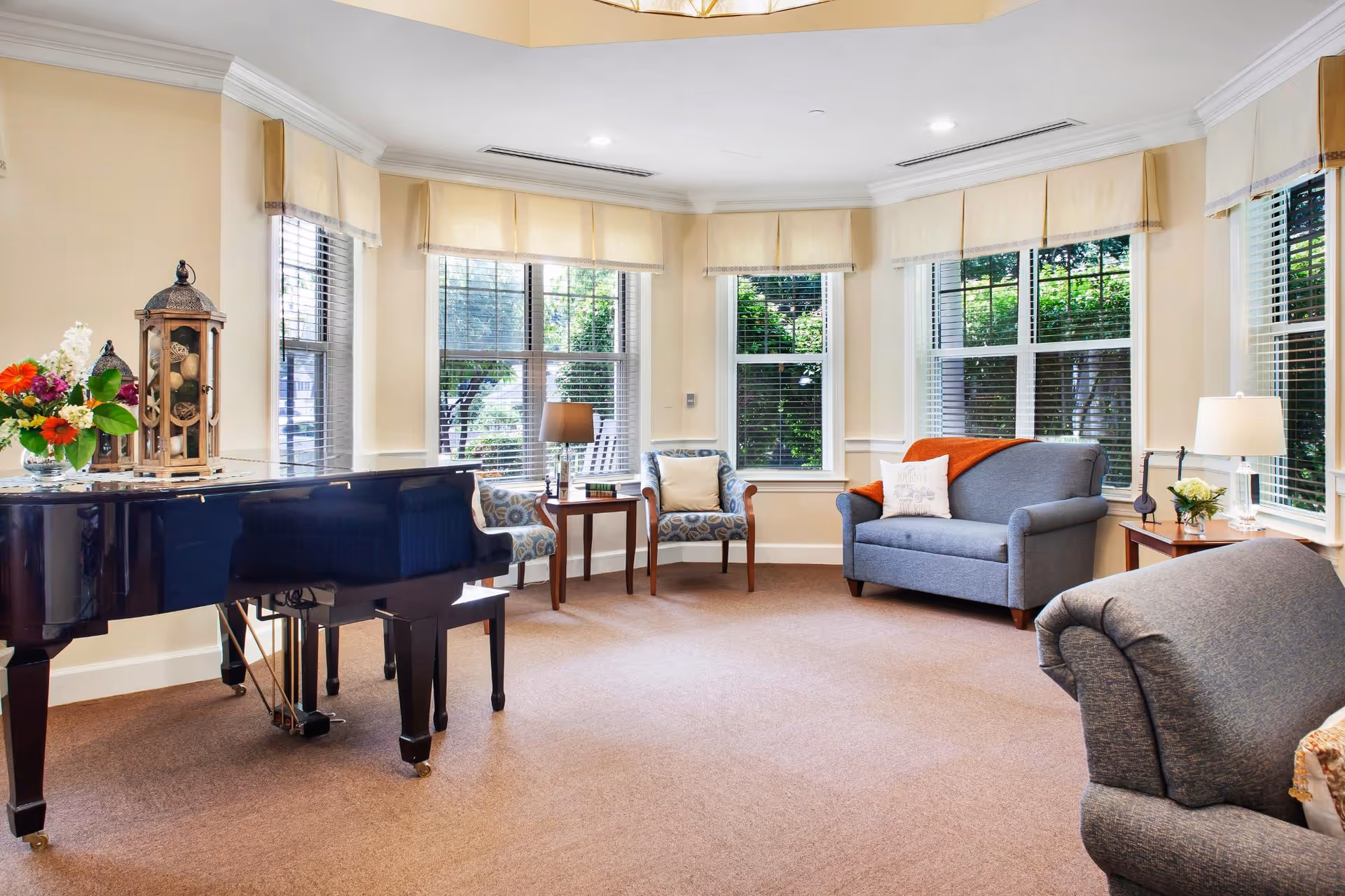 A bright and cozy living room with large windows letting in natural light. The room features a black grand piano with a floral arrangement on top, two patterned armchairs with a small wooden table and lamp between them, a gray loveseat with an orange throw blanket and a white pillow, and a gray armchair in the foreground. The walls are painted a soft cream color and the floor is carpeted in beige.