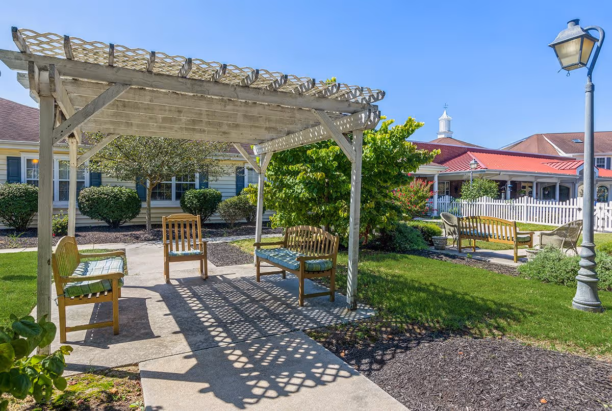 Outdoor seating area at Milford Place featuring wooden benches and chairs under a wooden pergola, surrounded by green grass, bushes, and a white picket fence with buildings in the background under a clear blue sky.