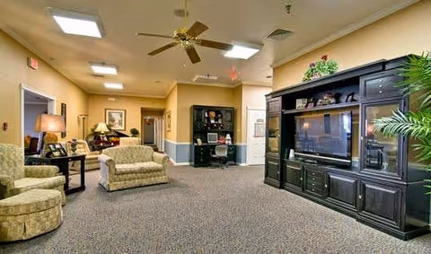 A spacious living room area in a senior living facility with beige walls and carpeted floor. The room features a large dark wood entertainment center with a flat-screen TV, a green patterned loveseat, a matching armchair, a side table with a lamp, and a ceiling fan with lights. There are framed pictures on the walls and a potted plant near the entertainment center.