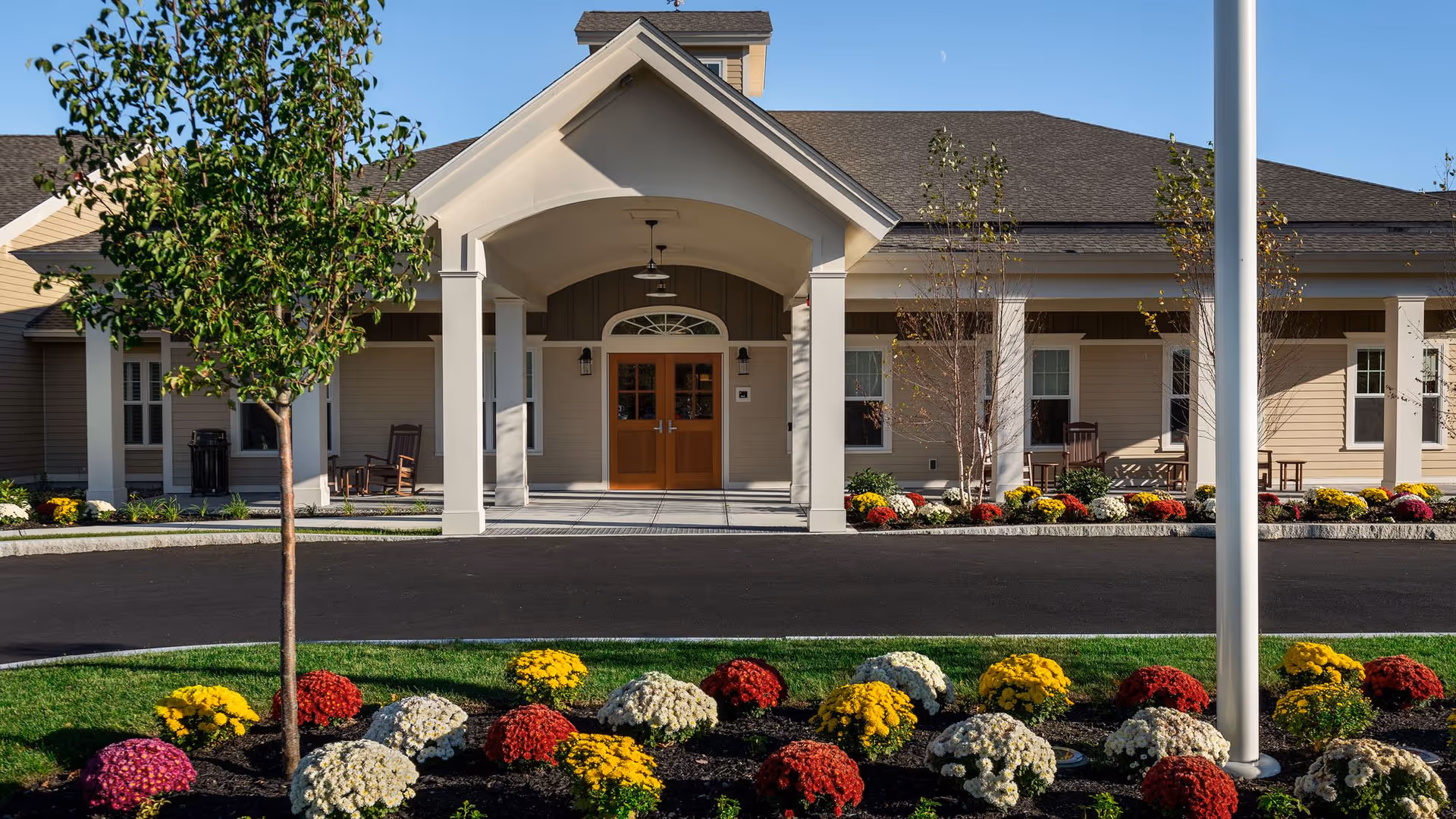 Front entrance of a senior living facility with a covered porch, double wooden doors, and a landscaped garden featuring colorful flowers and small trees under a clear blue sky.