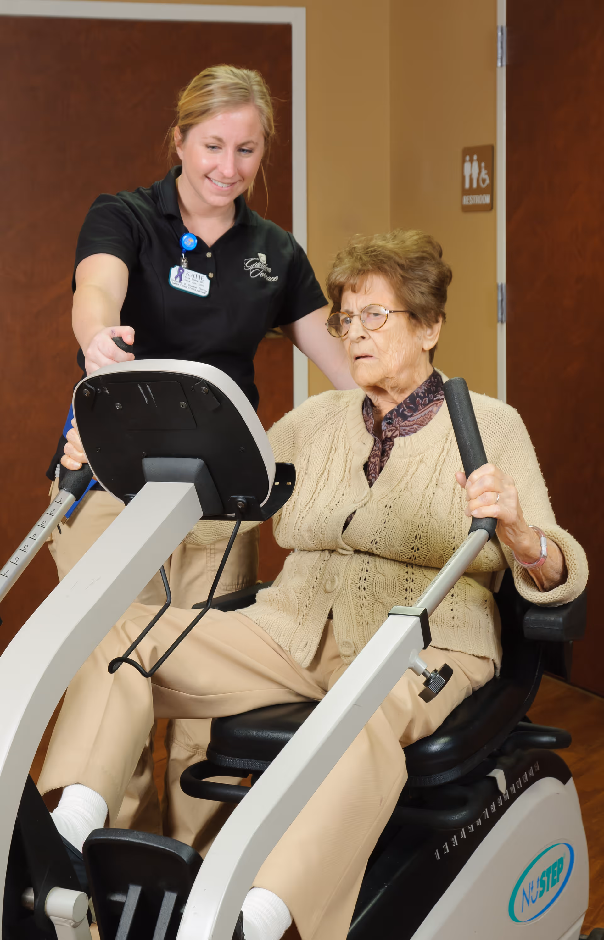 An elderly woman using a NuStep exercise machine with assistance from a smiling female caregiver wearing a Garden Terrace uniform in an indoor setting.