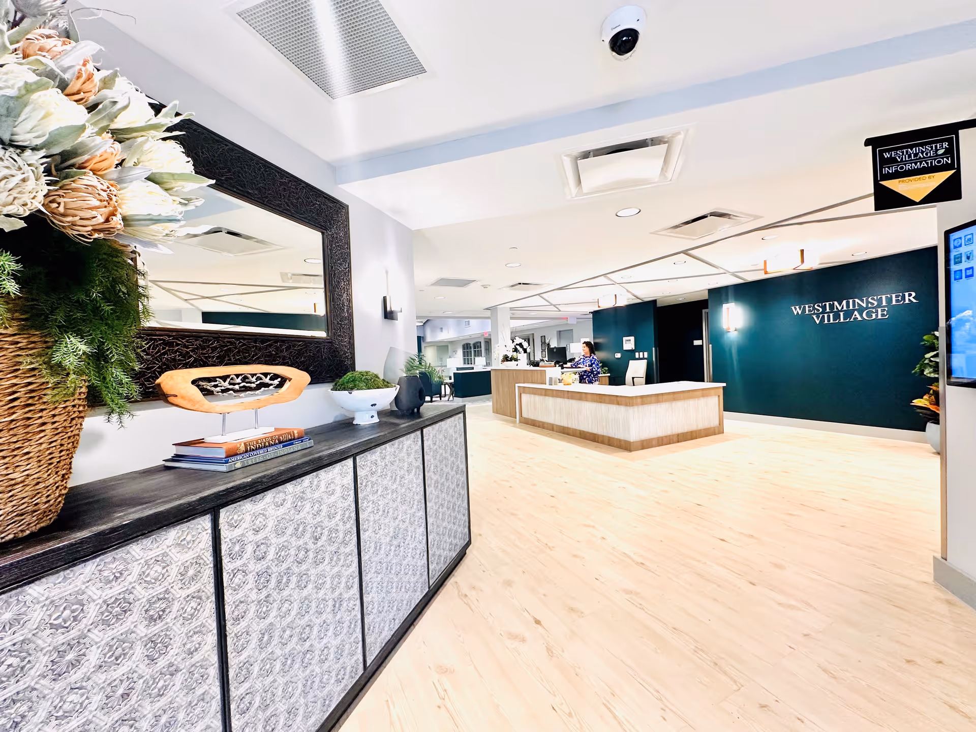 Bright and spacious reception area of Westminster Village with a wooden floor, a large decorative mirror above a cabinet with plants and books, and a reception desk where a staff member is seated. The walls are painted in white and dark green with the Westminster Village logo displayed prominently.