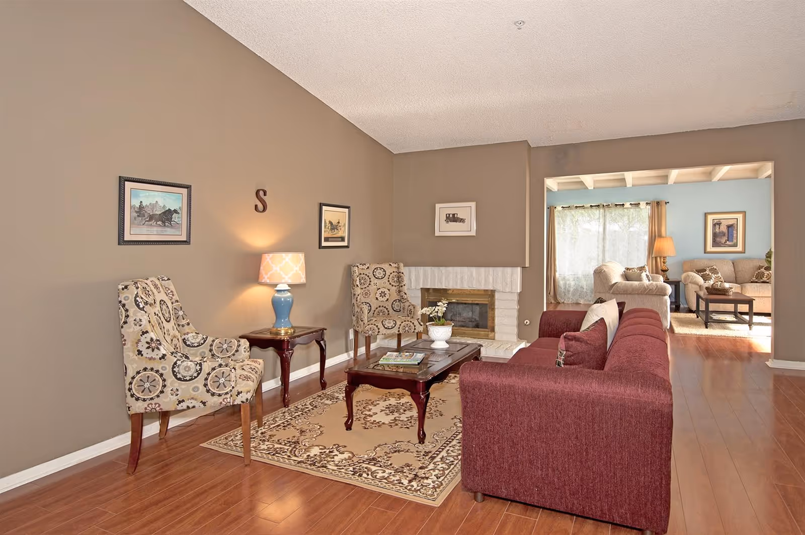 A cozy living room with a maroon sofa, two patterned armchairs, a wooden coffee table on a decorative rug, and a small side table with a blue lamp. The walls are painted beige with framed artwork and a letter 'S' hanging. There is a white brick fireplace and an adjoining room with additional seating and large windows with curtains.