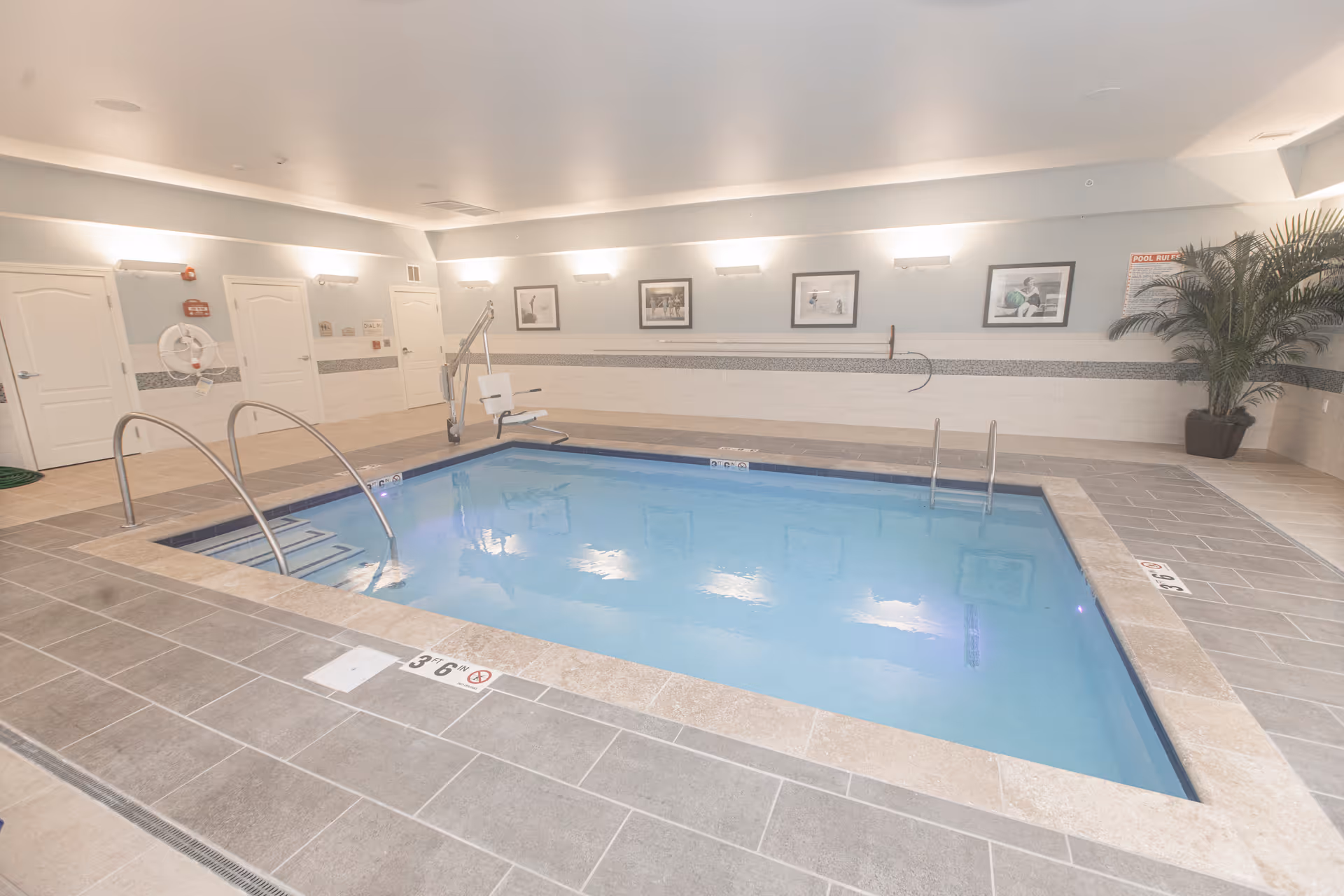 Indoor swimming pool with clear blue water surrounded by tiled flooring. The pool area has metal handrails and a pool lift chair for accessibility. The walls are light-colored with framed pictures and a large potted plant in the corner. Several closed doors and safety equipment like a life ring are visible on one side.