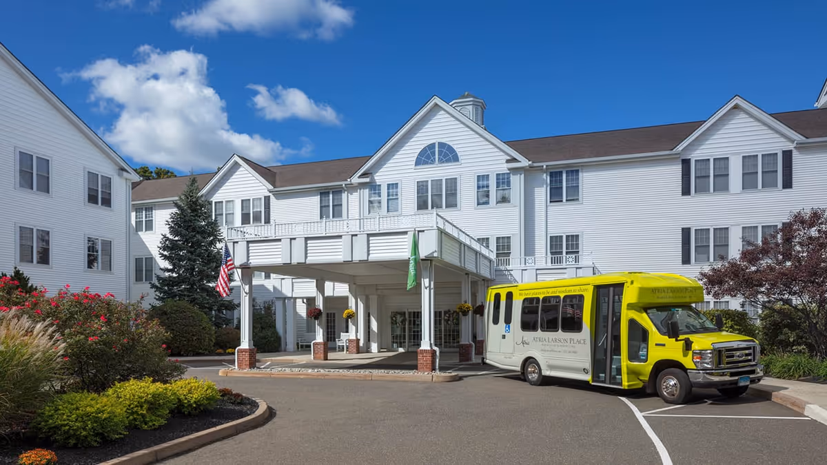 Exterior view of Atria Larson Place, a large white multi-story senior living facility with a covered entrance. A yellow and white shuttle bus with the facility's name is parked in front. The building is surrounded by landscaping with bushes, flowers, and trees under a blue sky with some clouds.