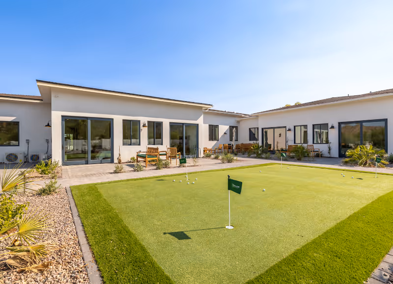 Sunlit courtyard with a small putting green, patio chairs and tables, and a one-story white building with large sliding glass doors.