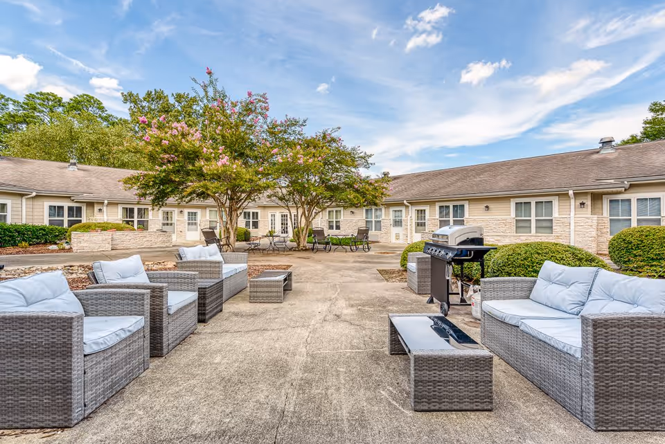 Outdoor courtyard area at Chesapeake Place Senior Living featuring cushioned wicker seating, a barbecue grill, trees with pink flowers, and a surrounding single-story building under a partly cloudy sky.
