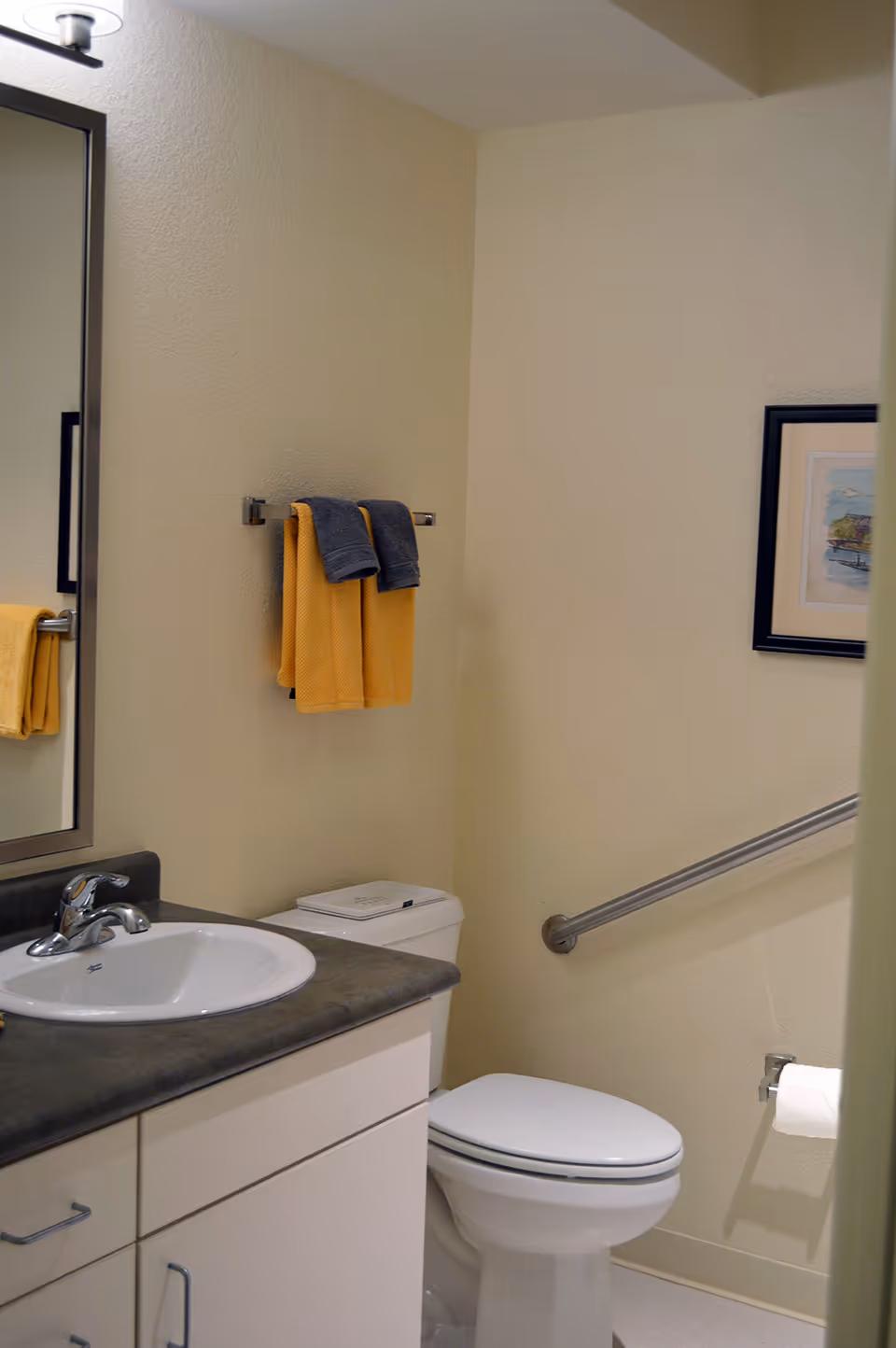 A clean bathroom featuring a white sink with a silver faucet set in a dark countertop, a white toilet with a closed lid, a metal grab bar on the wall next to the toilet, a towel rack holding two yellow towels and two gray towels, a framed picture on the wall, and a mirror above the sink.