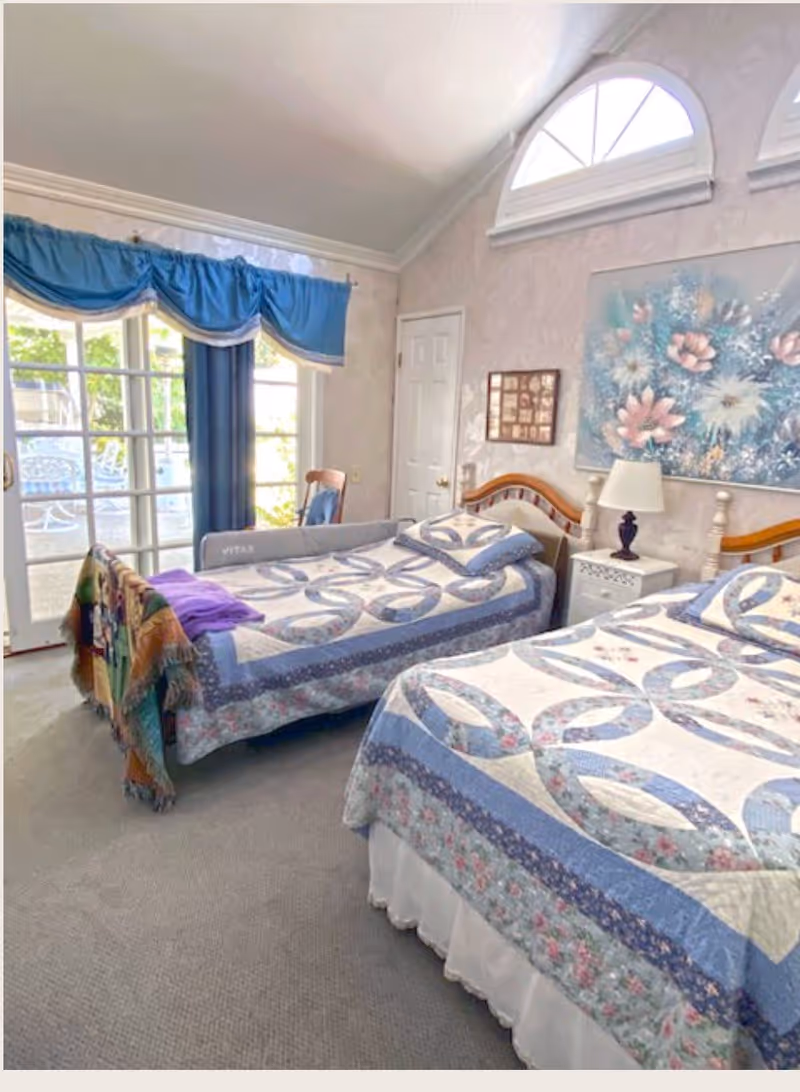 A bright bedroom with two twin beds covered in matching blue and white patterned quilts. There is a nightstand with a lamp between the beds, a floral painting on the wall, and a window with blue curtains letting in natural light. A door and a chair are visible near the window.
