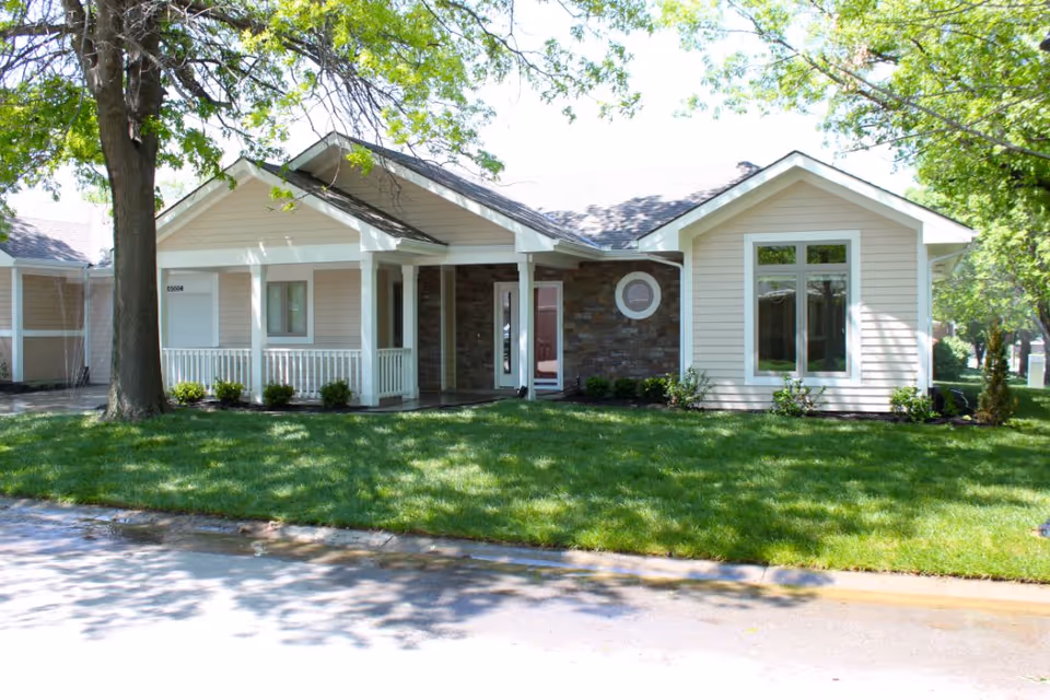 Front exterior view of a single-story residential building with beige siding, white trim, a covered porch with white railings, and a well-maintained green lawn with trees providing shade.